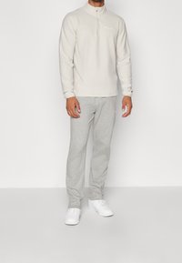 Muž v béžovém pulovru s čtvrtinovým zipem, světle šedých teplácích a bílých teniskách stojící proti jednoduchému bílému pozadí.