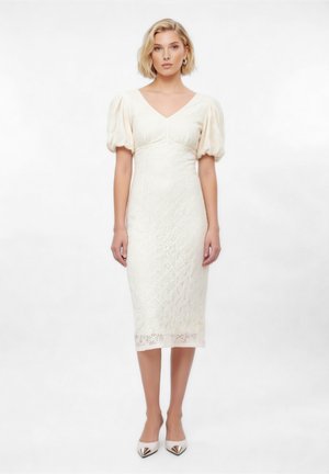 Kobieta stojąca w białej sukience midi z koronki z bufkami na rękawach i dekoltem w serek, nosząca białe szpilki z zaostrzonym noskiem na jednolitym tle.