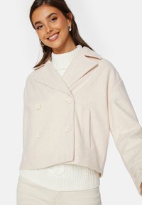 Světle růžová zkrácená bunda s dvojitými knoflíky, klopovém límci a boční kapsou. Texturovaná látka navrstvená na bílém pleteném svetru.