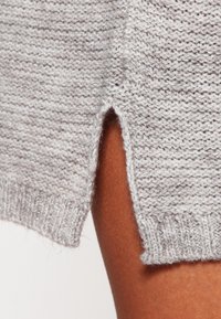 Ljusgrå stickad tröja med texturerad tyg, som har en sidreva och ribbad kant. Närbild som betonar stickat mönster.