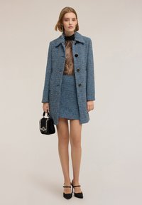 Modrý tvídový kabát s velkými černými knoflíky, v kombinaci s odpovídající sukní. Černý průhledný top pod ním, držící malou černou kabelku.