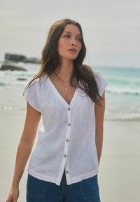 Chemise en lin blanche avec des manches courtes type châle, un décolleté en V et cinq boutons sur le devant ; associée à un short en jean bleu.