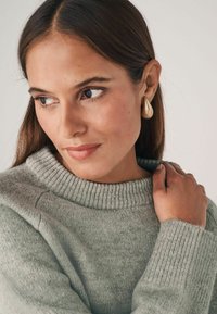 Jasnoszary dzianinowy sweter z prążkowanym kołnierzem i mankietami. Modelka ma długie brązowe włosy i nosi złote kolczyki w kształcie kropli.