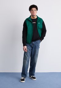 Chaleco acolchado verde sobre una camiseta negra de manga larga, combinado con unos pantalones vaqueros anchos azules y sneakers negros. Diseño simple sin patrones visibles.