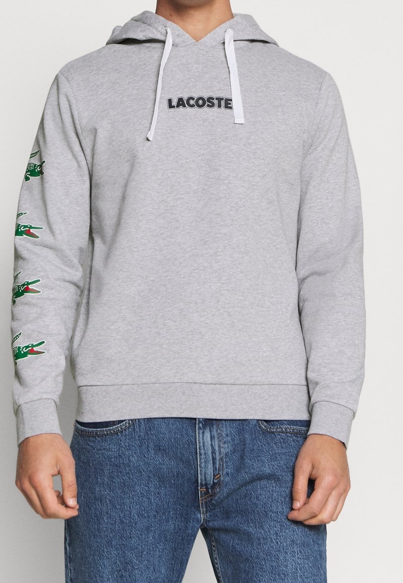 Personne portant un sweat à capuche Lacoste gris clair avec des cordons blancs et des logos crocodile verts le long de la manche gauche, associé à un jean bleu.