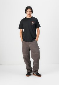 Sort bomulds T-shirt med grafisk print foran, parret med løstsiddende, grå denimbukser med synlige syninger og et sidelabel. Sorte sneakers.