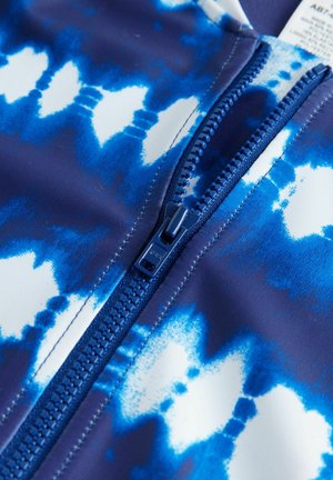 Rash guard azul y blanco con un patrón de tie-dye, un zipper central en azul y una textura de tela suave y elástica.
