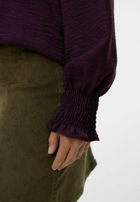 Mano appoggiata di lato, mentre indossa una blusa lunga a maniche scure viola con polsini arricciati e una gonna di corduroy verde oliva.