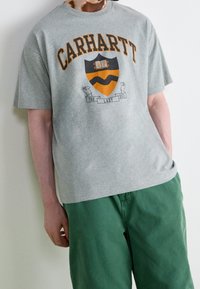 Graues Kurzarm-T-Shirt mit "CARHARTT" in Orange, ausgestattet mit einem Schildgrafik und einem Bannerdesign "THE LAZY DUCK" in der Mitte.