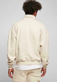 Uomo con capelli ricci che indossa un felpa color crema e pantaloni abbinati, in piedi di spalle contro uno sfondo grigio uniforme.