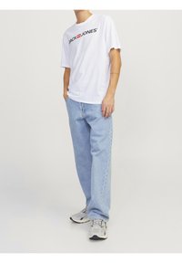 Camiseta blanca de manga corta con el logo "JACK & JONES", combinada con jeans rectos de mezclilla azul claro y zapatillas grises con detalles en negro.