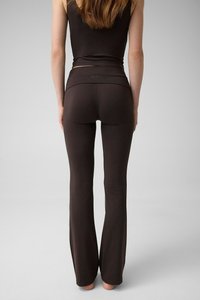 Femme debout pieds nus, portant un haut noir moulant sans manches et un pantalon noir évasé, vue de dos sur un fond gris uni.
