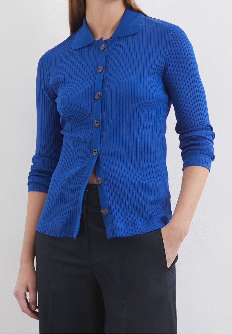 Femme portant un cardigan bleu côtelé ajusté boutonné avec des manches trois-quarts et un pantalon noir, une main dans la poche, sur un fond uni.