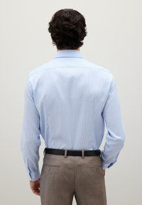 Uomo con capelli ricci scuri che indossa una camicia azzurra chiara infilata in pantaloni beige con una cintura nera, in piedi di spalle.