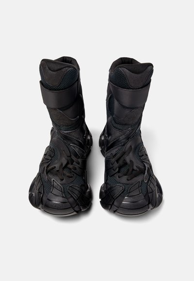 Svarta ankelstövlar med en texturerad ovansida, kardborreband och en kraftig sula med en intrikat design. Materialet verkar hållbart och flexibelt.