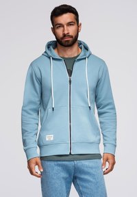 Jasnoniebieska bluza z kapturem na zamek błyskawiczny, z białymi sznurkami, przednimi kieszeniami i małą metką z logo. Nosi się ją na ciemnej koszulce, w komplecie z dżinsami.