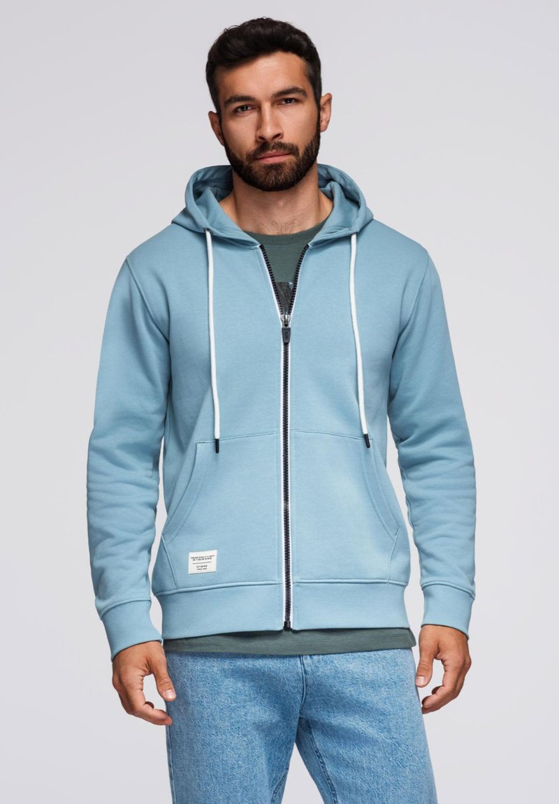 Jasnoniebieska bluza z kapturem na zamek błyskawiczny, z białymi sznurkami, przednimi kieszeniami i małą metką z logo. Nosi się ją na ciemnej koszulce, w komplecie z dżinsami.