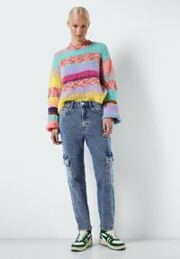 Suéter de punto multicolor con rayas en rosa, azul, amarillo y verde, combinado con unos pantalones cargo de mezclilla azul claro y zapatillas verdes y blancas.