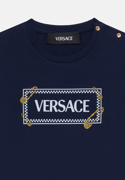 Tamsiai mėlyna medvilninė marškinėlių su balta "VERSACE" logotipu, suporuotu su languota rėmeliu ir auksiniais saugos segtukais dekoracijoms netoli kaklo.