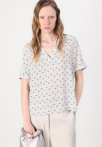 Blouse à manches courtes en V, en tissu léger, présentant un motif géométrique noir et blanc. Associée à un pantalon beige clair et un sac argenté.