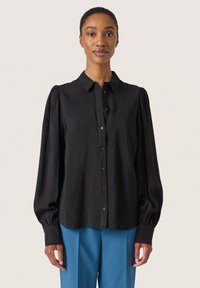 Bluză lungă neagră cu mâneci lungi, cu nasturi în față, umeri bufanți și textură moale. Asortată cu o pereche de pantaloni albaștri, creând un aspect contrastant.