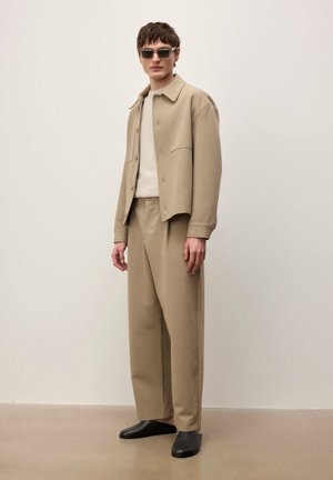 Mužský model v béžovom saku a nohaviciach, bielej košeli, čiernych slip-on topánkach a tmavých slnečných okuliaroch na jednoduchom svetlom pozadí.