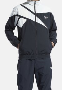 Person som bär en svart och vit Reebok-träningsdräkt med händerna i fickorna, med en framdragbar dragkedja samt elastiska ärmslut och midja.