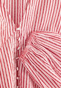 Hvidt stof med smalle, lodrette røde striber, med stofbeklædte knapper og elastisk, rynket manchet på lang ærme.