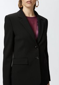 Crni blazer odijelo s dva prednja džepa i tri gumba. Tkanina izgleda glatko s laganim sjajem, naglašavajući njegovu strukturiranu dizajn.