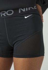 Black Nike sports shorts with a gray logo waistband, featuring a mesh panel on the side and smooth texture. Designed for performance and comfort.