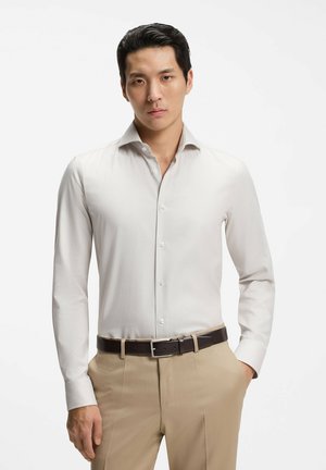 Ung mand iført en hvid skjorte med knapper stukket ned i beige bukser med et mørkebrunt bælte, står med den ene hånd i lommen.