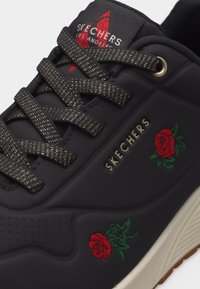 Baskets Skechers noires avec lacets dorés, broderie d'une rose rouge et logo Skechers sur le côté et la languette.