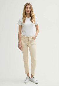 Witte t-shirt gecombineerd met lichtbeige, slim-fit jeans met een vijfknopengulp. Gedragen met witte sneakers tegen een lichte achtergrond.