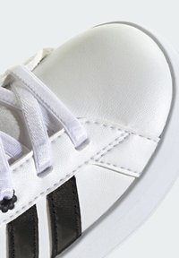 Pointe de basket blanche avec lacets blancs et deux bandes verticales noires sur le côté, montrant une couture détaillée et une texture.