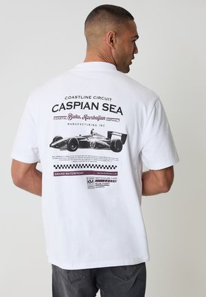 Mężczyzna w białej koszulce z czarno-bordowym nadrukiem wyścigowego samochodu oraz napisem "Morze Kaspijskie Baku, Azerbejdżan" z tyłu.