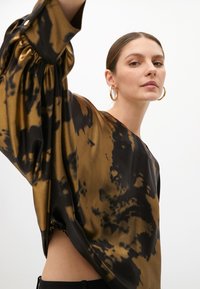 Blusa con effetto tie-dye marrone e nero, con maniche larghe e dettaglio di arricciatura laterale. Realizzata in tessuto lucido, presenta un ampio scollo.
