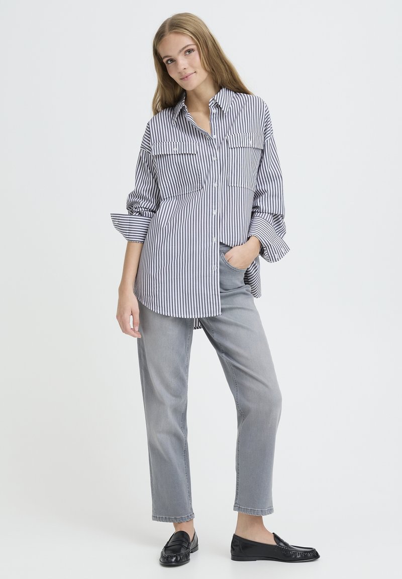 Striped button-up shirt with two chest pockets, loose fit, and rolled sleeves. Paired with grey pants and black loafers on a plain background.