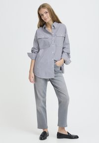 Chemise à rayures avec deux poches poitrine, coupe ample et manches retroussées. Associée à un pantalon gris et des mocassins noirs sur un fond uni.