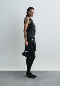 Czarna kamizelka o fakturze z przednimi kieszeniami i perełkowymi zdobieniami, zestawiona z plisowaną czarną spódnicą. Czarne rajstopy i szpilki w szpic. Mała torebka.