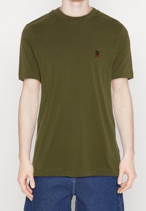 Man draagt een olijfgroen T-shirt met korte mouwen en een klein geborduurd logo op de linkerborst, gecombineerd met blauwe spijkerbroek.