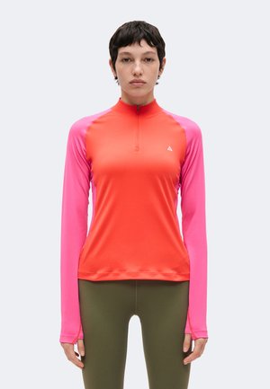 Kvinde med kort mørkt hår iført en langærmet atletisk top med orange krop og pink ærmer, parret med olivengrønne leggings.