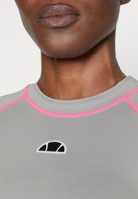 Šedý atletický top s kulatým výstřihem, zdobený růžovým šitím a malým černo-bílým logem na hrudi. Hladká textura.