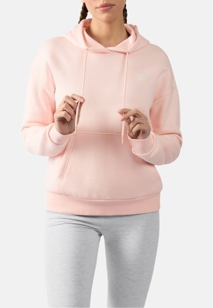 Vrouw draagt een lichtroze hoodie en lichtgrijze legging, houdt de koordjes van de hoodie vast tegen een effen achtergrond.
