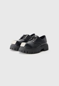 Scarpe in pelle nera con suola in gomma spessa, dotate di un accento metallico quadrato sulla punta e lacci rotondi per la chiusura.