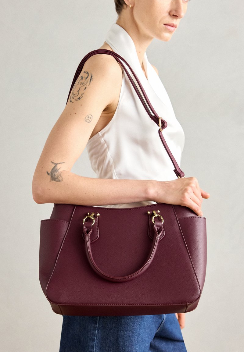 Bolso de piel burdeos con dos asas superiores y una correa de hombro desmontable. Superficie texturizada con acentos de herrajes en tono dorado.