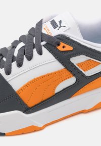 Puma-tennarit, joissa on harmaa, valkoinen ja oranssi värimaailma, nahka- ja verkkomateriaaleja, tekstuuripinnat sekä näkyvä brändäys.