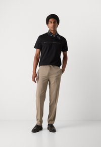 Tricou negru cu logo în relief, pantaloni bej cu talie elastică și pantofi negri. O bandană cu un model este legată la gât.