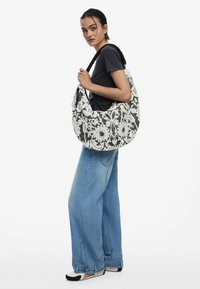 Schwarz-weiße, mit Blumenmuster verzierte Stofftasche mit geschwungener Form, strukturiertem Oberflächen und einem schwarzen geflochtenen Tragegurt, die über einem schlichten T-Shirt getragen wird.