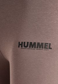 Tejido de ropa deportiva marrón con una textura suave. Presenta un logotipo impreso en negro "HUMMEL" y "SPORTSWEAR SINCE 1923."