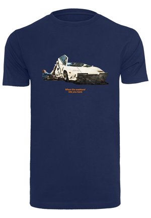 Osoba leżąca na ziemi obok białego sportowego samochodu z otwartymi drzwiami, na granatowej koszulce z napisem "Kiedy weekend uderza cię mocno".